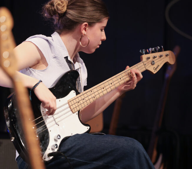 A female student sits and plays a black electric guitar on stage during a concert