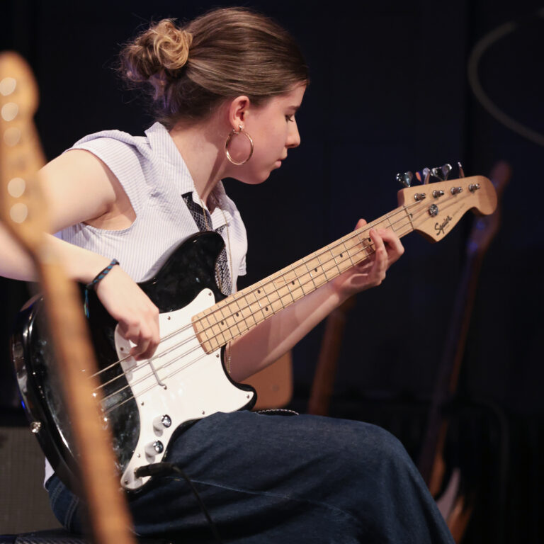 A female student sits and plays a black electric guitar on stage during a concert
