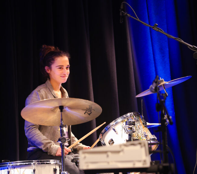 A female student sits behind a drum kit on stage