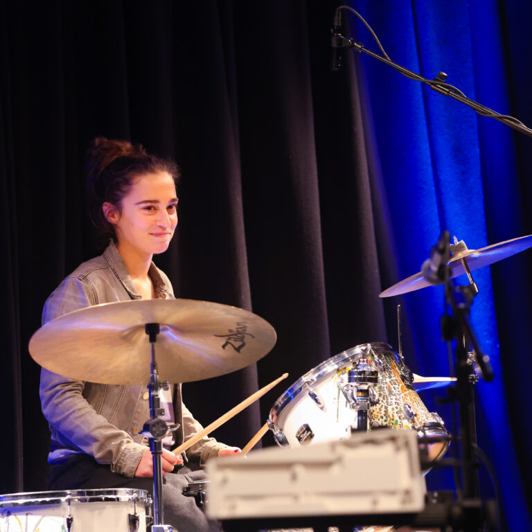 A female student sits behind a drum kit on stage