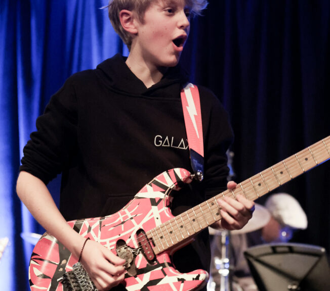 A male student wearing a black hoody plats a patterned electric guitar on stage