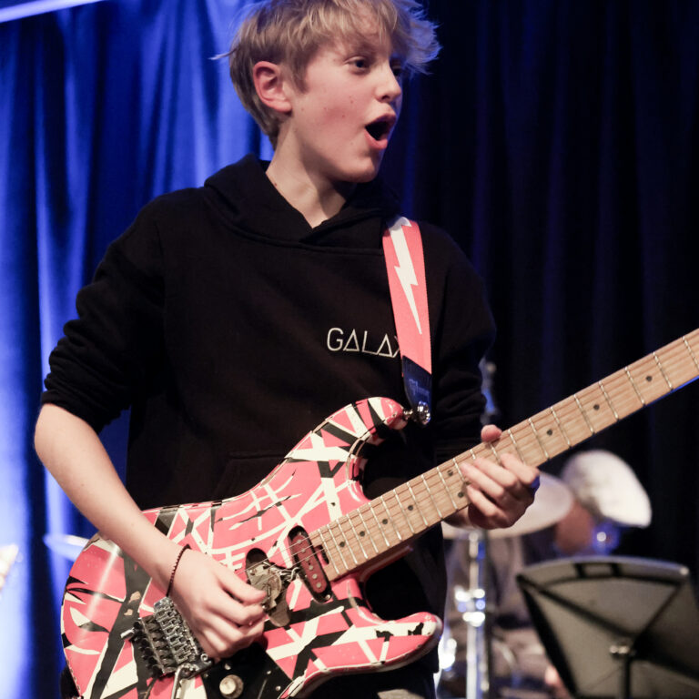 A male student wearing a black hoody plats a patterned electric guitar on stage