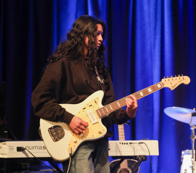 A female student in a black hoody plays a white electric guitar on stage during a concert performance