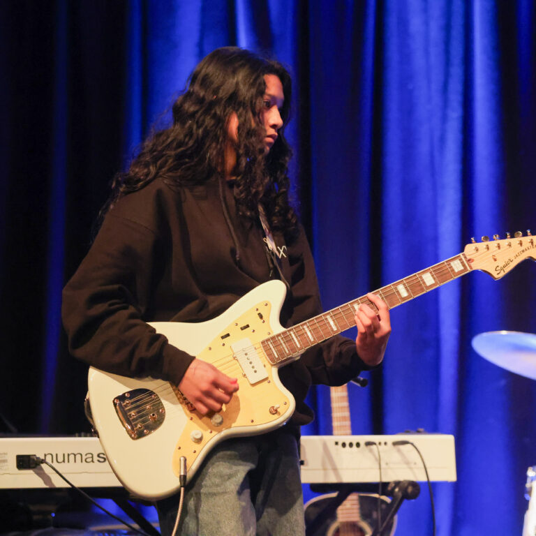 A female student in a black hoody plays a white electric guitar on stage during a concert performance