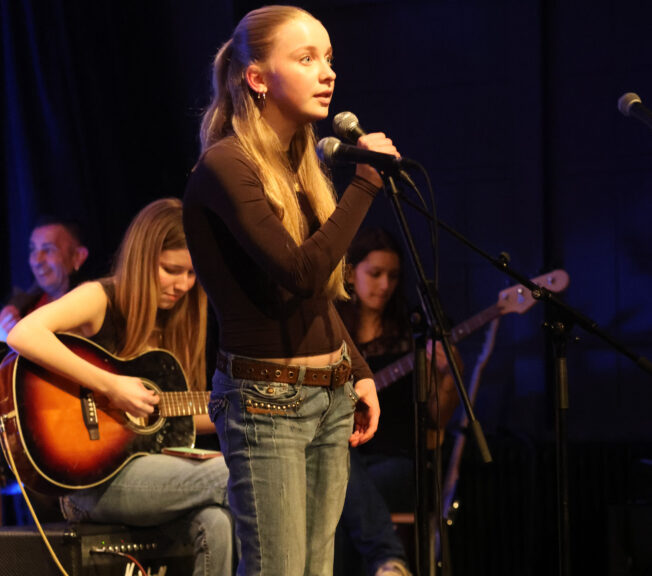 A female student holds a microphone on stage, with musicians sat behind her