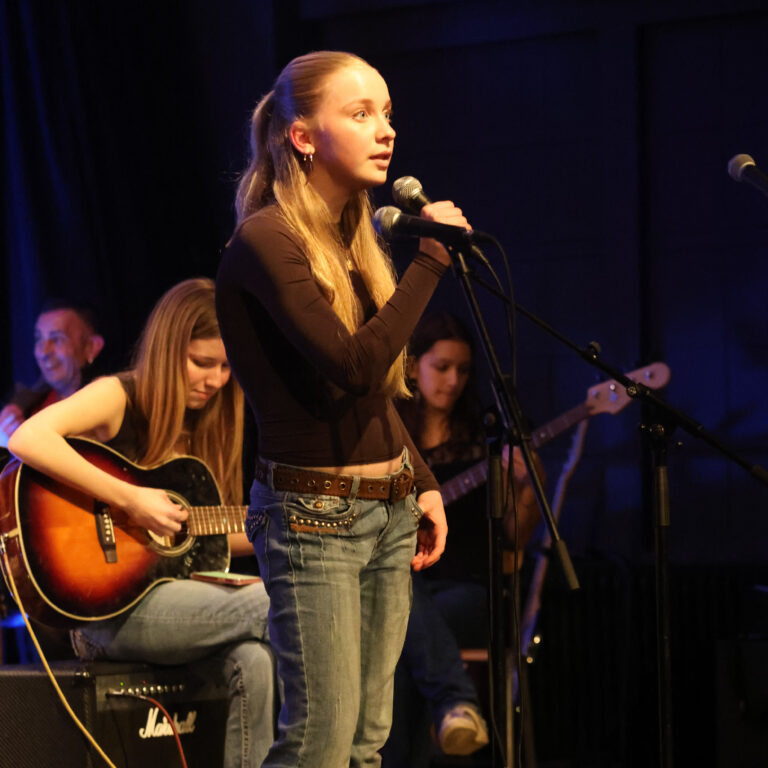A female student holds a microphone on stage, with musicians sat behind her