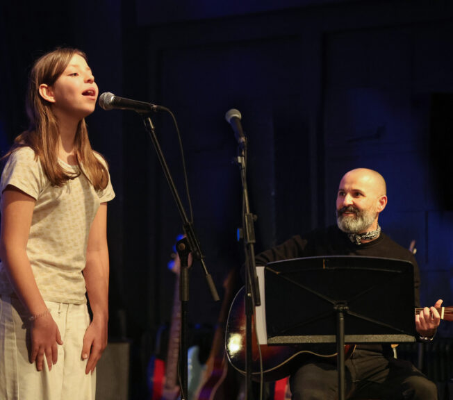A female student sings into a microphone on stage, accompanied by a male teacher on piano