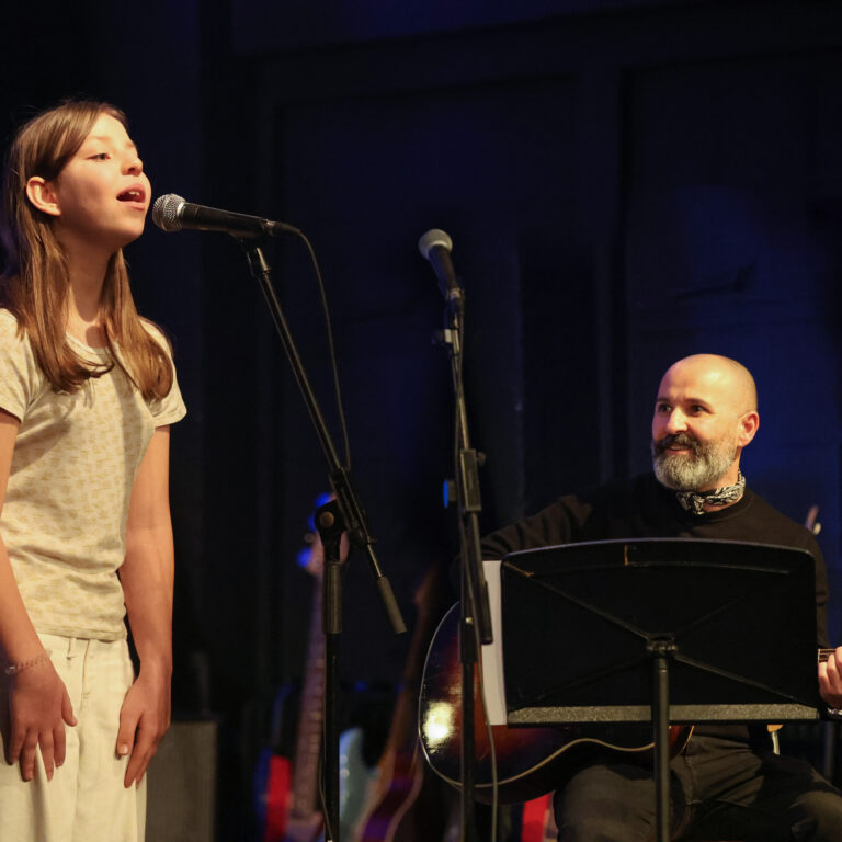 A female student sings into a microphone on stage, accompanied by a male teacher on piano
