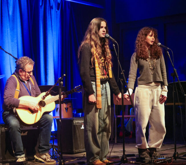 Two female students hold hands on stage whilst singing into microphones, accompanied by a male teacher on guitar