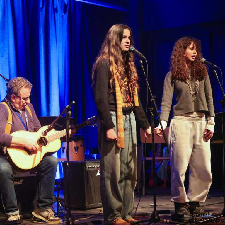 Two female students hold hands on stage whilst singing into microphones, accompanied by a male teacher on guitar