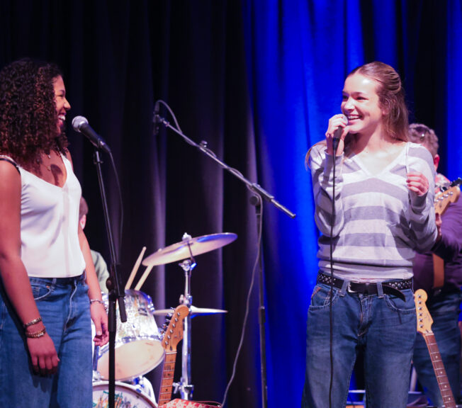 Two female students smile at each other on stage whilst singing