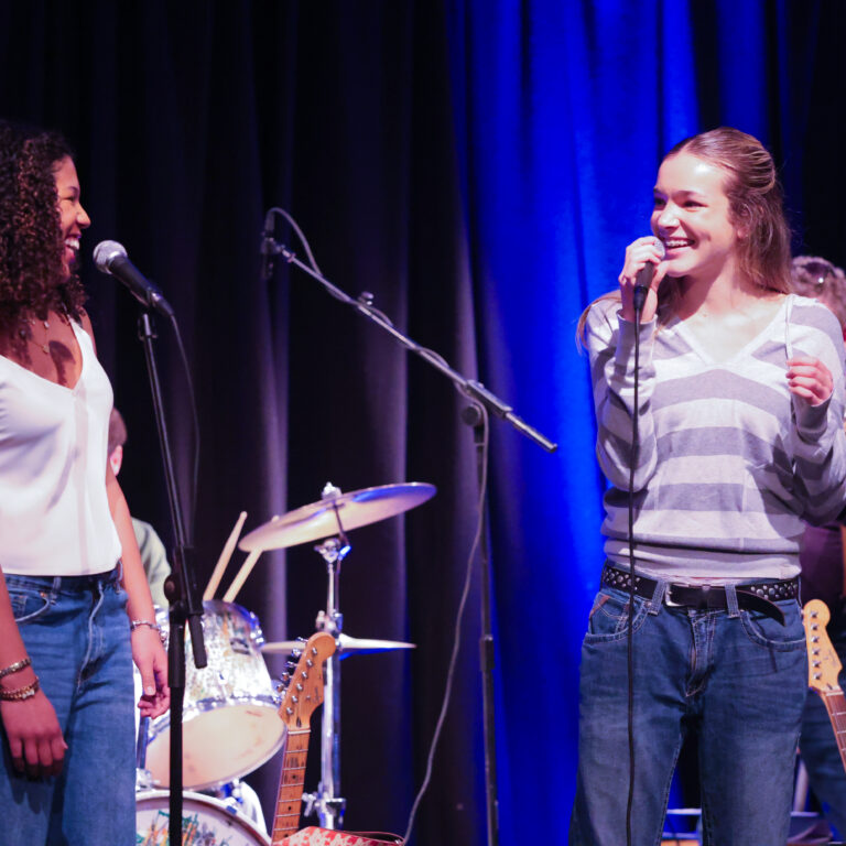 Two female students smile at each other on stage whilst singing