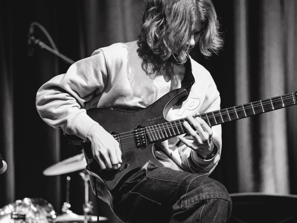 Black and white image of a male student with long hair playing an electric guitar
