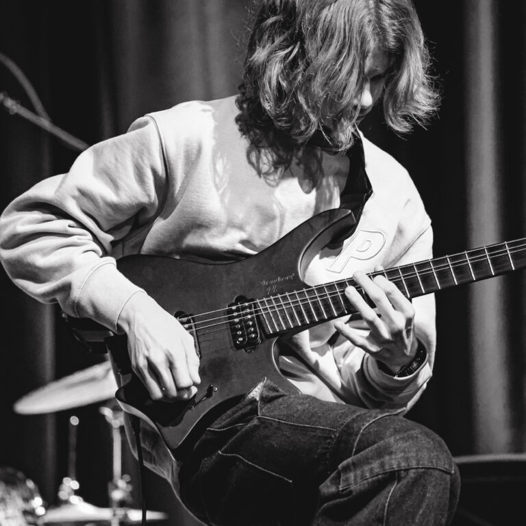 Black and white image of a male student with long hair playing an electric guitar
