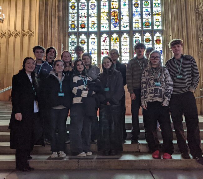 A group of Sixth Form students from The King Alfred School, North London, pose for a picture on stone steps in front of a grand stained glass window.