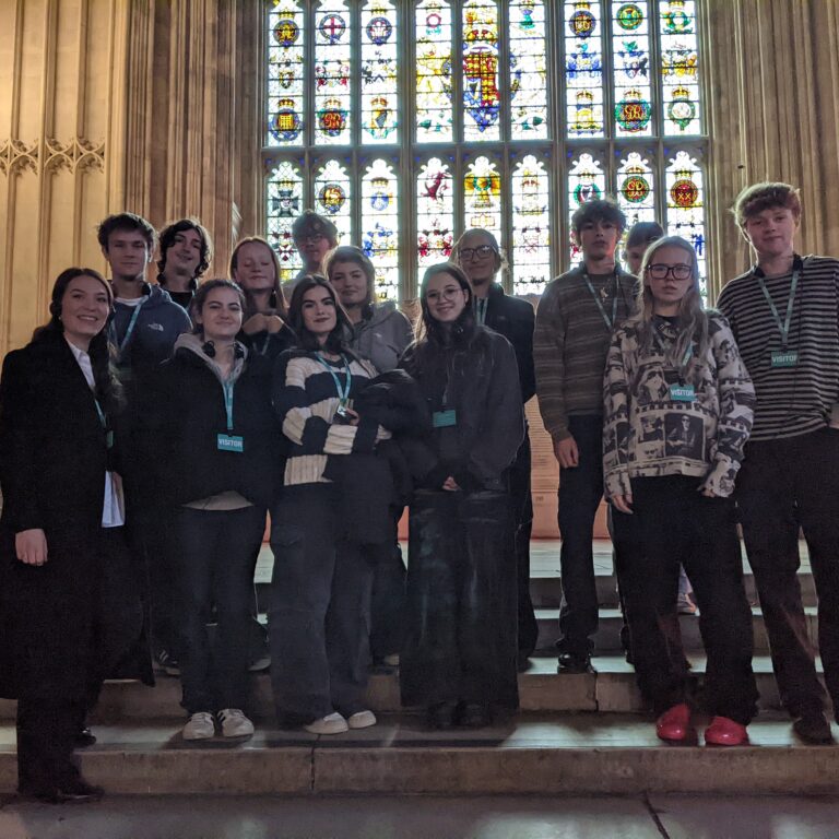 A group of Sixth Form students from The King Alfred School, North London, pose for a picture on stone steps in front of a grand stained glass window.