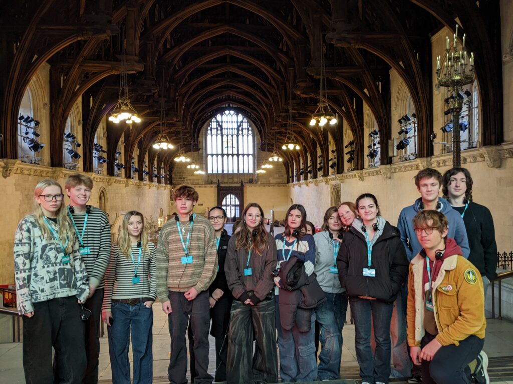 A line of Sixth Form students pose for a picture in a grand hall, featuring an arched wooden roof and stained glass windows.