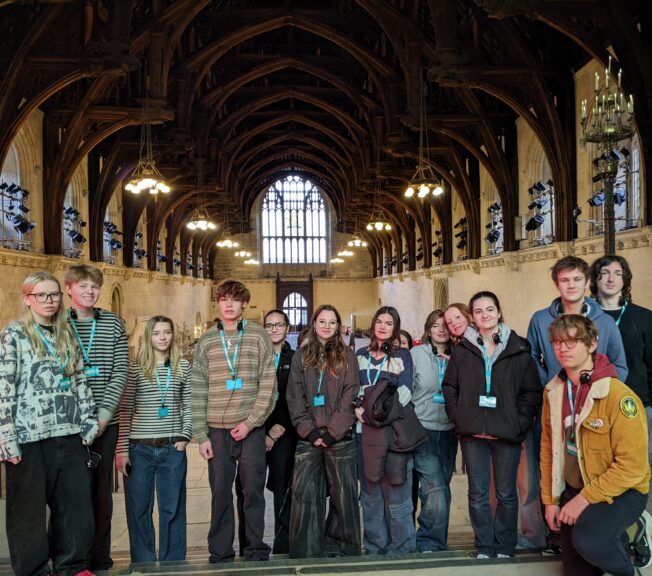 A line of Sixth Form students pose for a picture in a grand hall, featuring an arched wooden roof and stained glass windows.