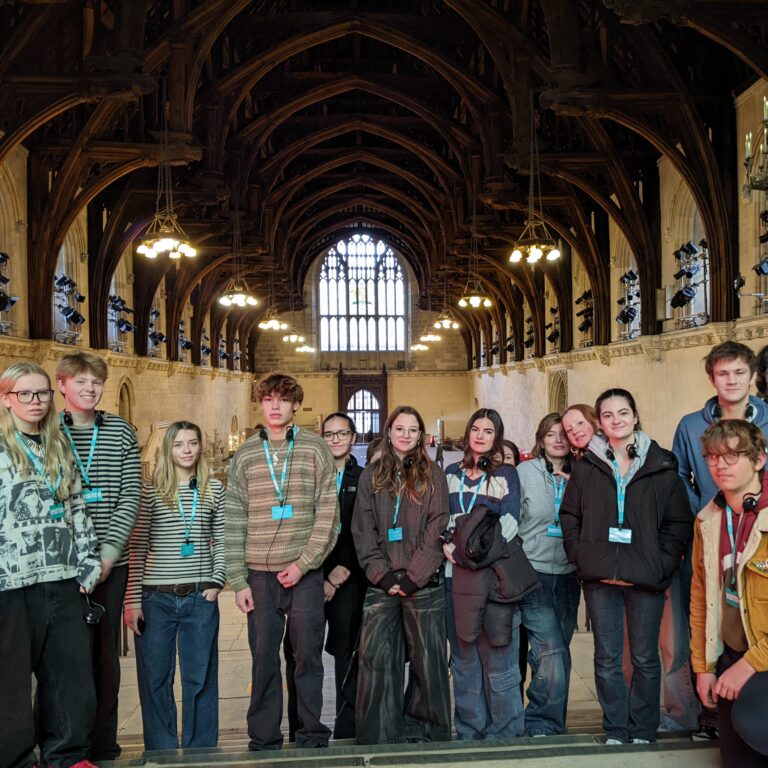 A line of Sixth Form students pose for a picture in a grand hall, featuring an arched wooden roof and stained glass windows.