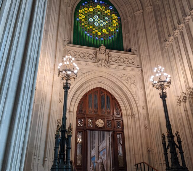 Ornate stone wall, wooden arched door, and stained glass window at the Houses of Parliament, UK. Ornate tall lamps flank stone steps.