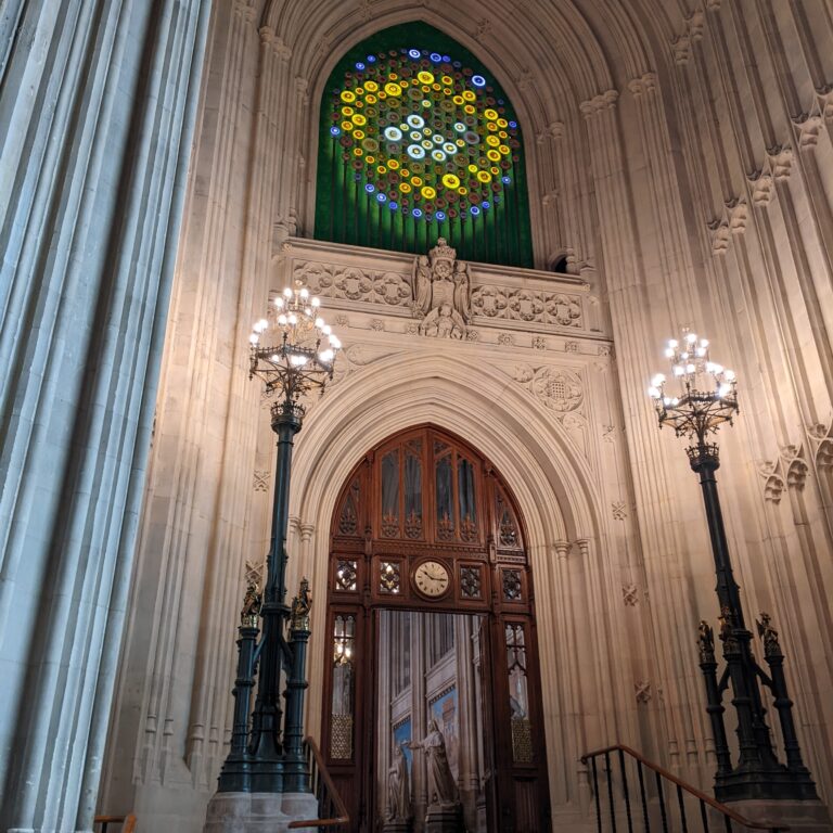 Ornate stone wall, wooden arched door, and stained glass window at the Houses of Parliament, UK. Ornate tall lamps flank stone steps.