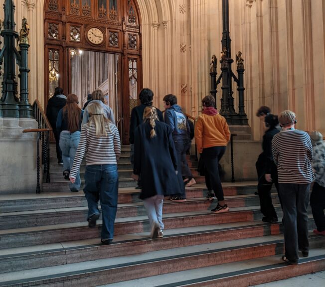 A group of Sixth Form students from The King Alfred School, North London, walk up stone steps to an ornate wooden door at the Houses of Parliament, UK