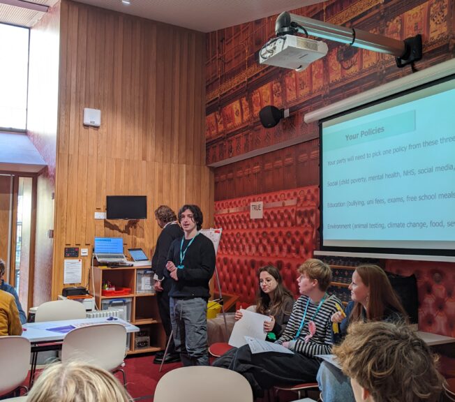 A male student stands in front of fellow students, presenting a slide of fictional government policies as part of a Politics trip to the Houses of Parliament