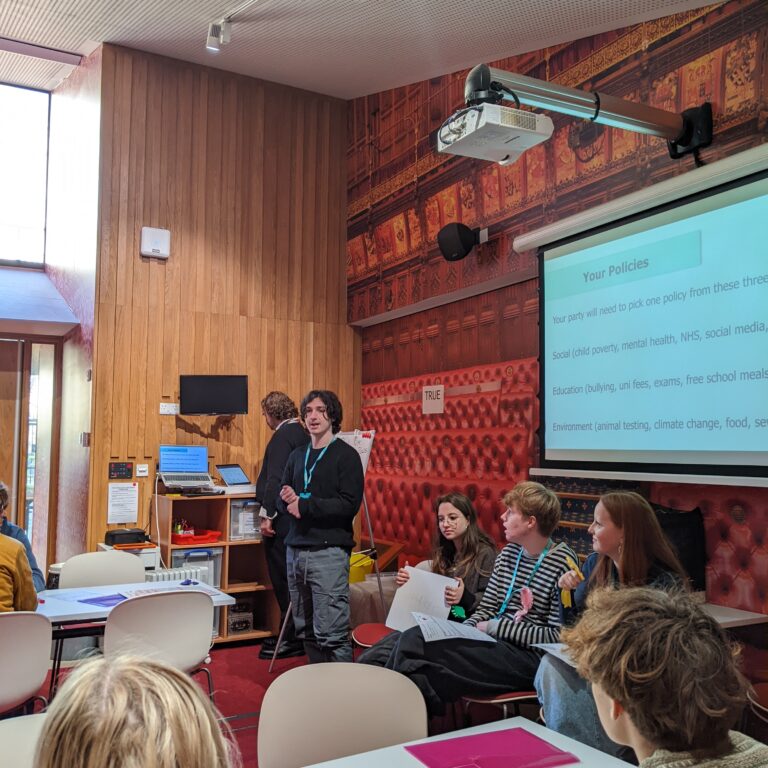 A male student stands in front of fellow students, presenting a slide of fictional government policies as part of a Politics trip to the Houses of Parliament