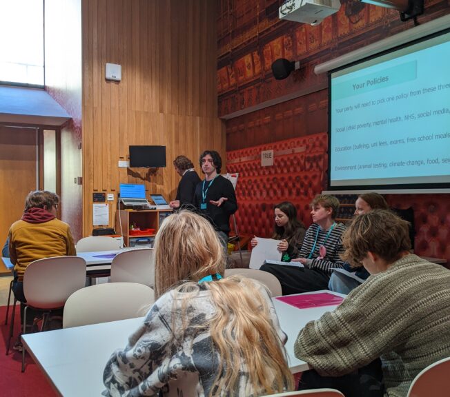 A male student stands in front of fellow students, presenting a slide of fictional government policies as part of a Politics trip to the Houses of Parliament