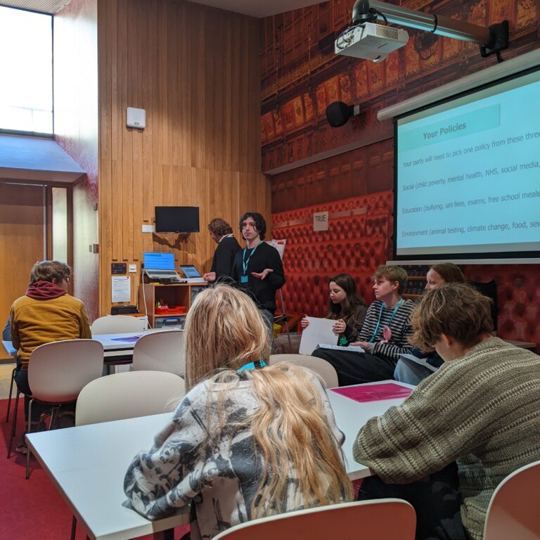 A male student stands in front of fellow students, presenting a slide of fictional government policies as part of a Politics trip to the Houses of Parliament