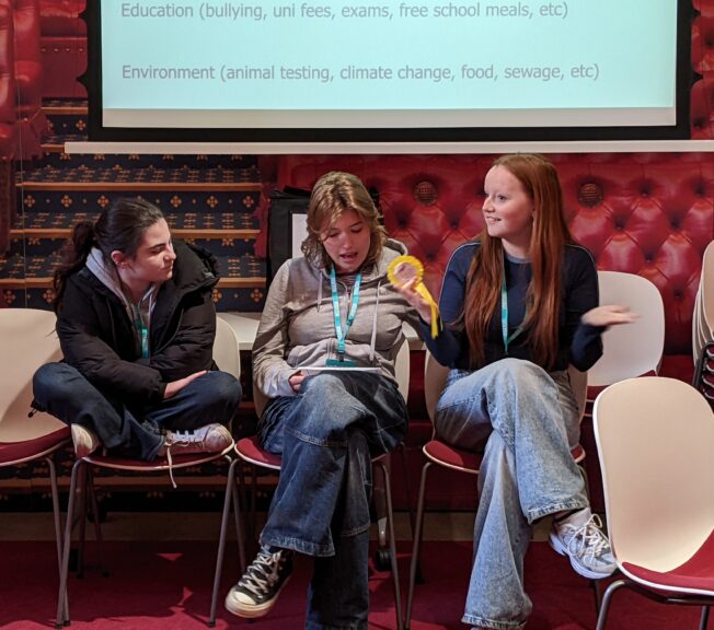 Three female students sit and talk animatedly in front of a projector screen and a House of Lords backdrop