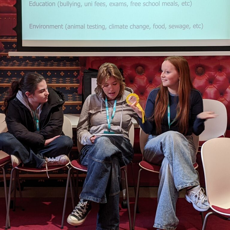 Three female students sit and talk animatedly in front of a projector screen and a House of Lords backdrop