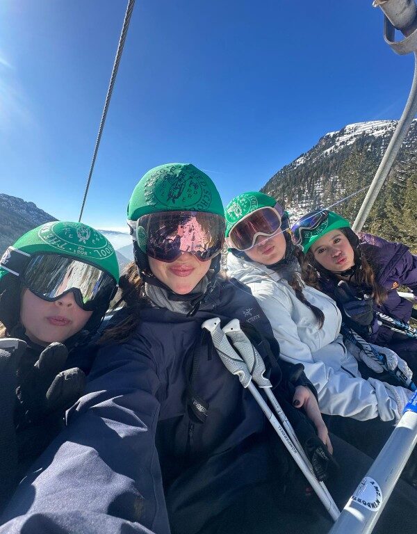 Four students wearing ski goggles and helmets pose on a ski lift, with alpine trees and a blue sky behind them