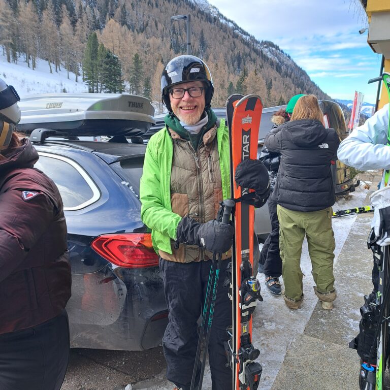 A male teacher from The King Alfred School holds his skis in front of a snowy mountain scene