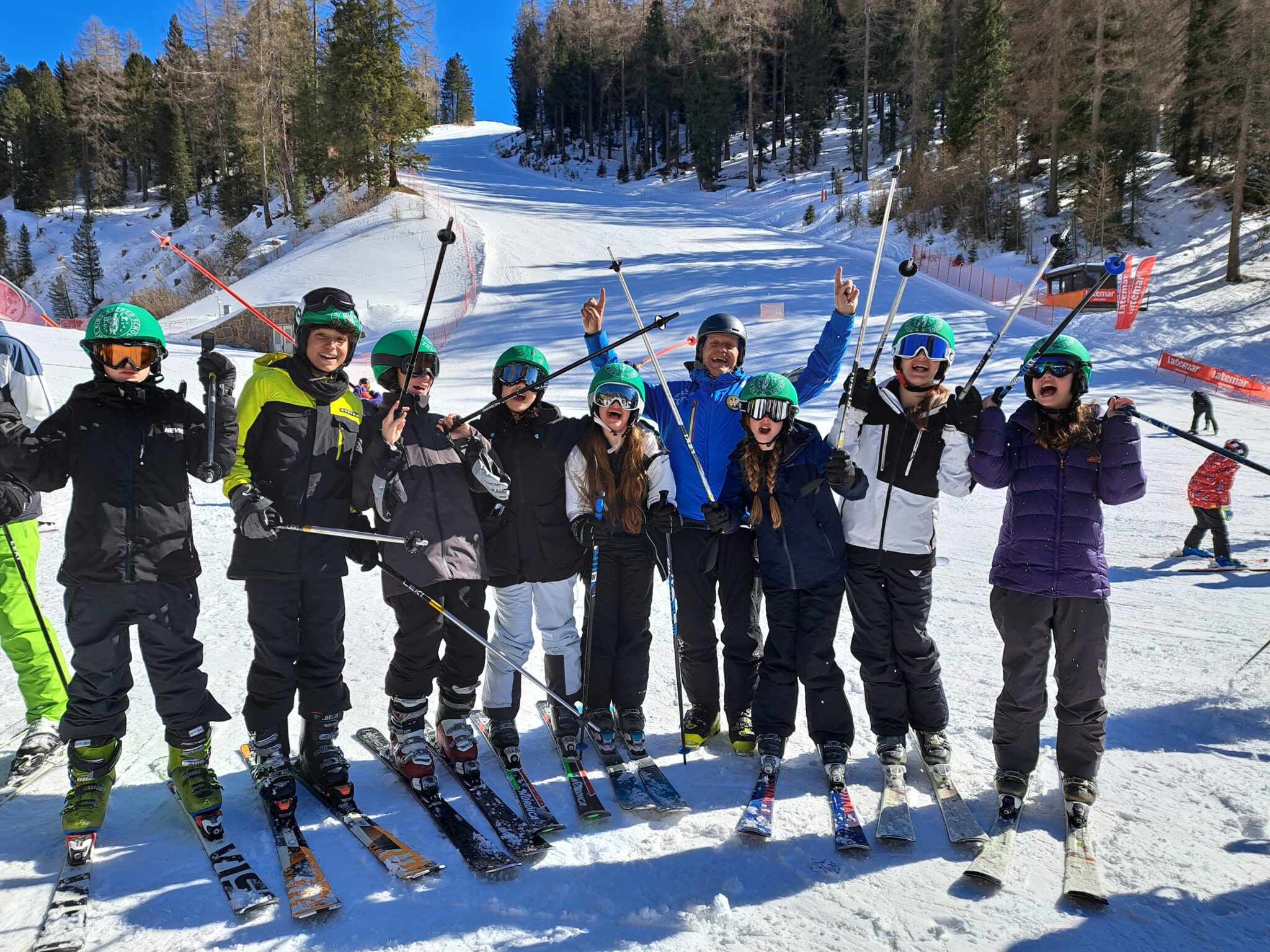Students from The King Alfred School and their ski instructor stand in a line on a snowy ski slope, raising their ski poles and cheering