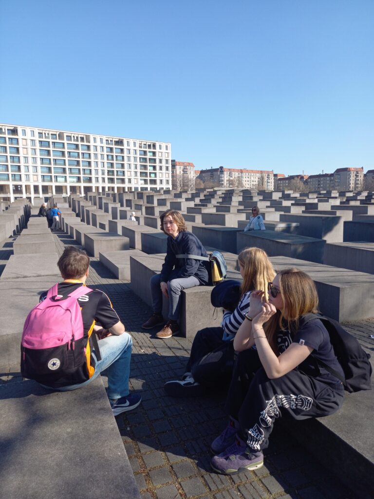 A group of five students sit outside on a concrete floor, with concrete blocks of different heights - a Holocaust memorial site in Berlin - behind them
