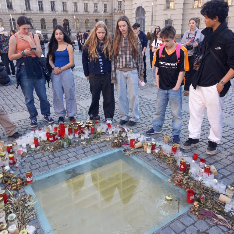 A group of students look at candles and flowers left in tribute on the ground of a square in Berlin