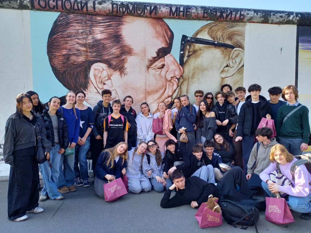 A large crowd of students stand and kneel in front of a section of the Berlin Wall, which depicts Soviet leader Leonid Brezhnev kissing East German leader Erich Honecker