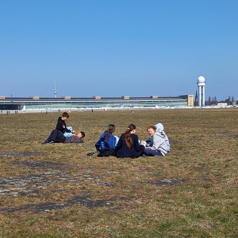 Four students sit on a vast open field in Berlin, with a low building and blue sky in the distance