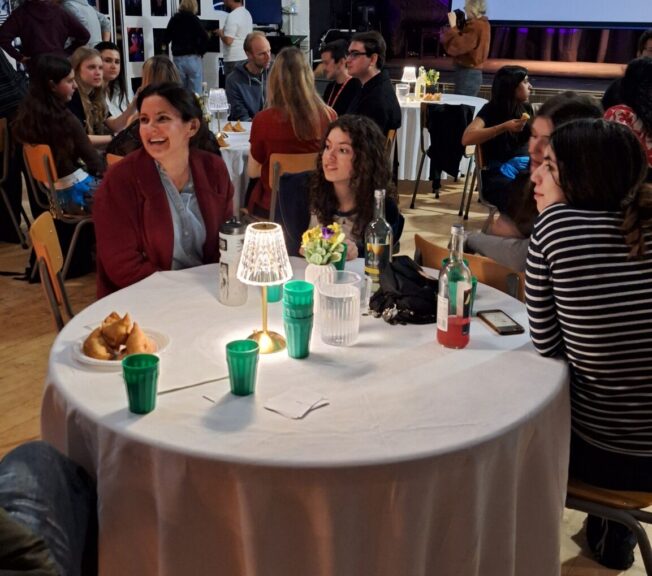 A parent and students sit at a round table in a school hall, laughing and talking