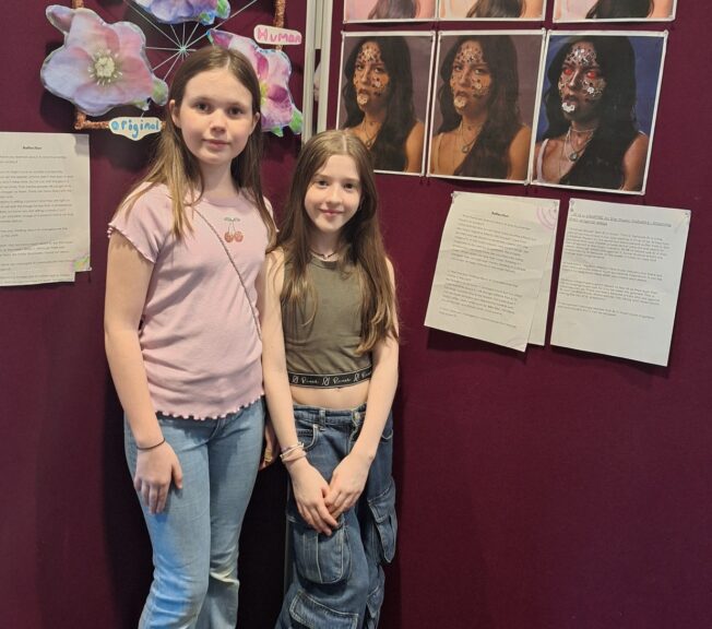 Two female students pose next to a display of their work on digital manipulation of art and music using AI