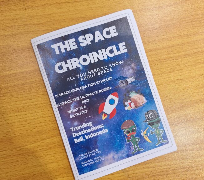 A student-produced magazine lies on a school table. It is titled 'The Space Chronicle' with the sub-header: 'Is space exploration ethical?'