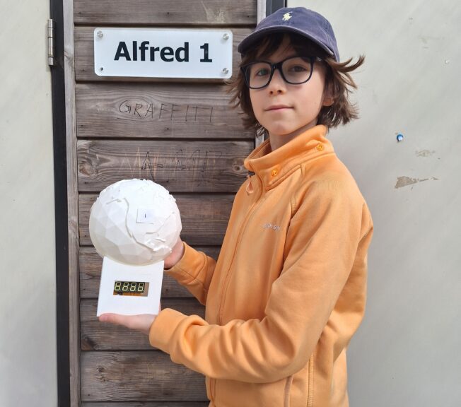 A male student wearing glasses and a cap poses in front of a school building holding a 3D printed model of the globe which he made