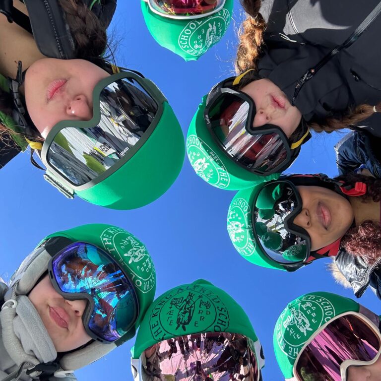 A ring of students wearing ski goggles and helmets bearing The King Alfred School logo look down at the camera from above, with a bright blue sky behind them