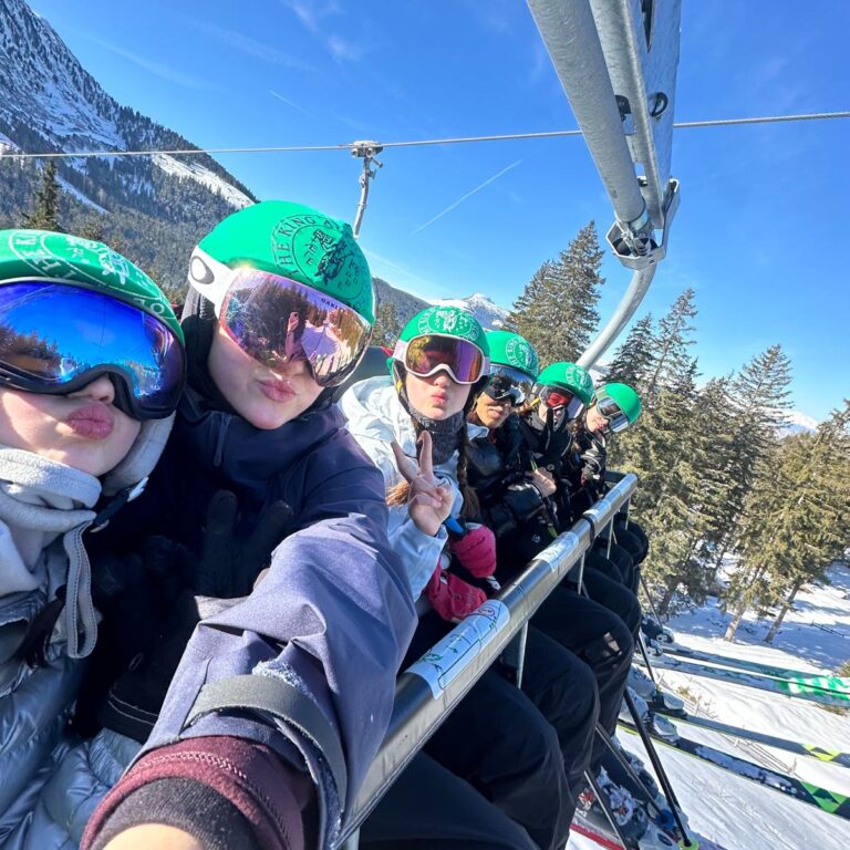 Students wearing ski goggles and helmets bearing The King Alfred School logo pose on a ski lift, with alpine trees and a blue sky behind them