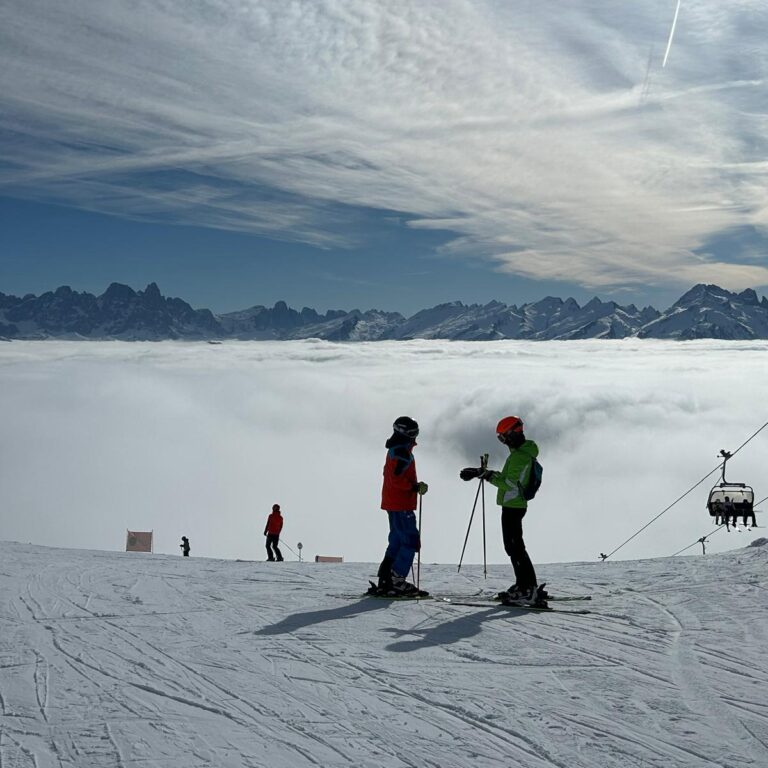 Two students on skis appear in shadow on top of a sunny, snowy mountain scene