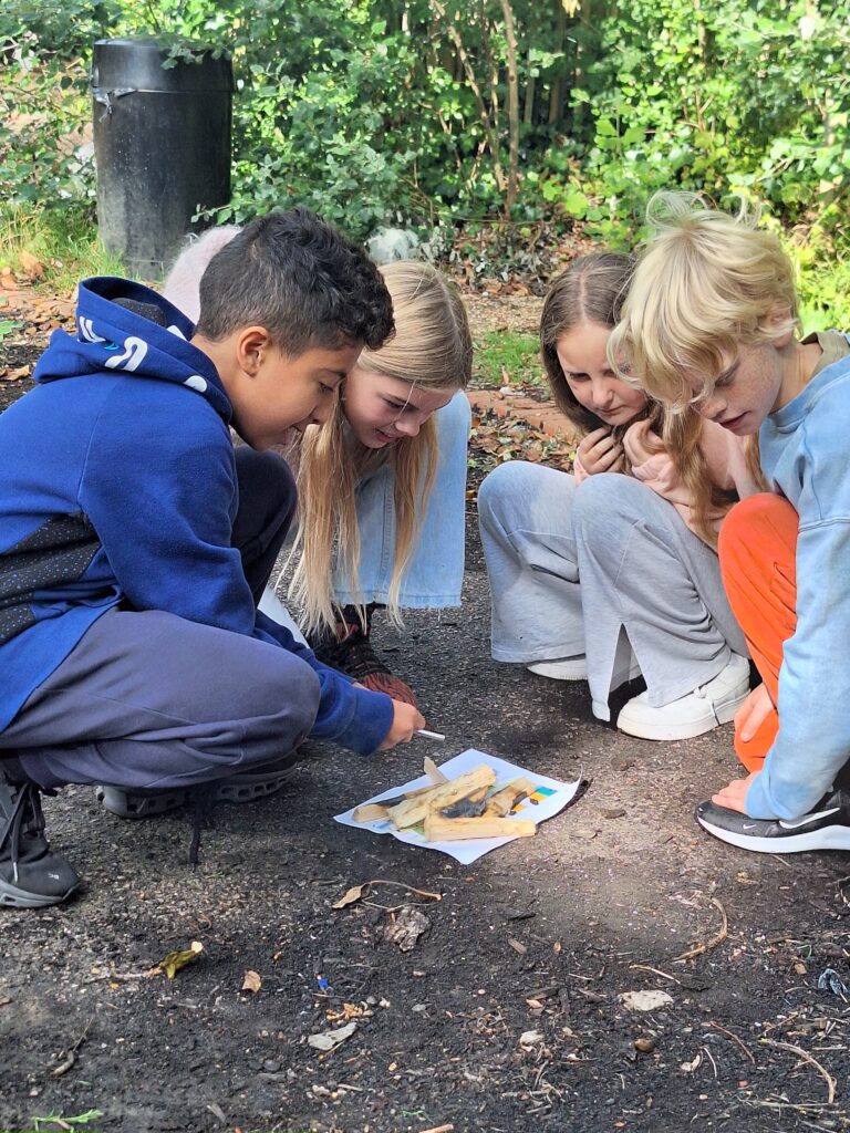 A group of young students crouch down close to the ground as one attempts to light a fire using a flint and steel