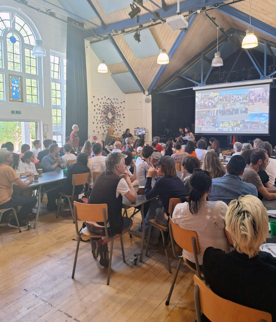 Students, teachers and parents in a school hall, with two students talking at a lectern and a selection of photos on a big screen