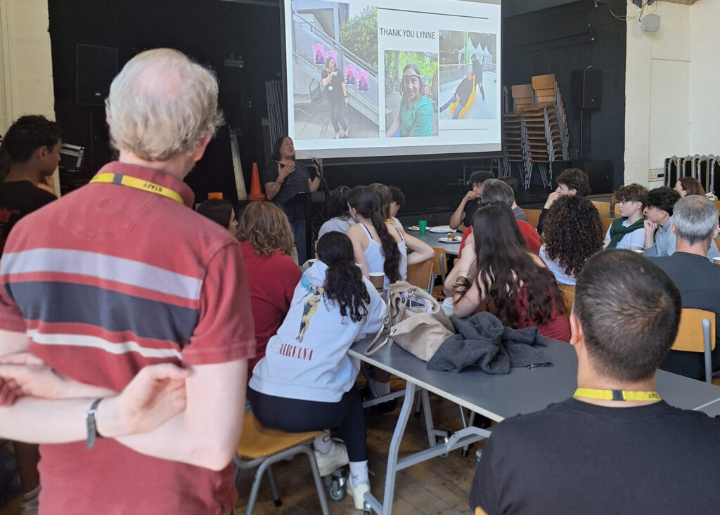 A teacher talks to a crowd of students and parents in a school hall