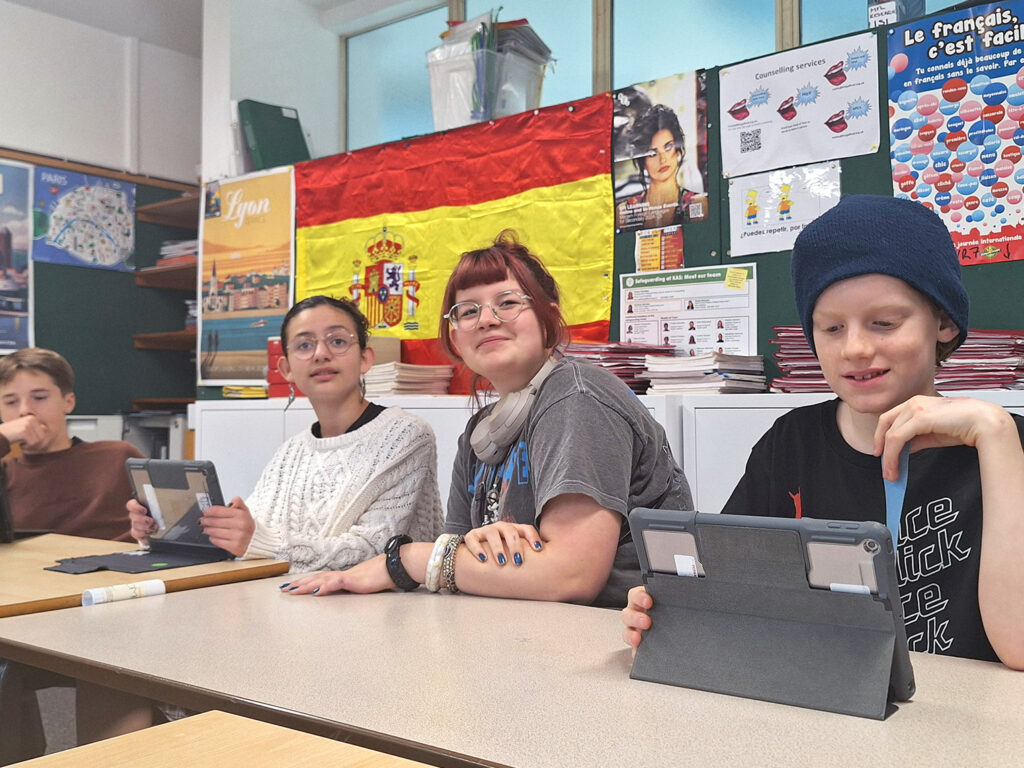 Two male and two female students sit at desks in a Modern Foreign Languages classroom, looking at iPads and the front of the room. There are displays of work and a large Spanish flag in the background.